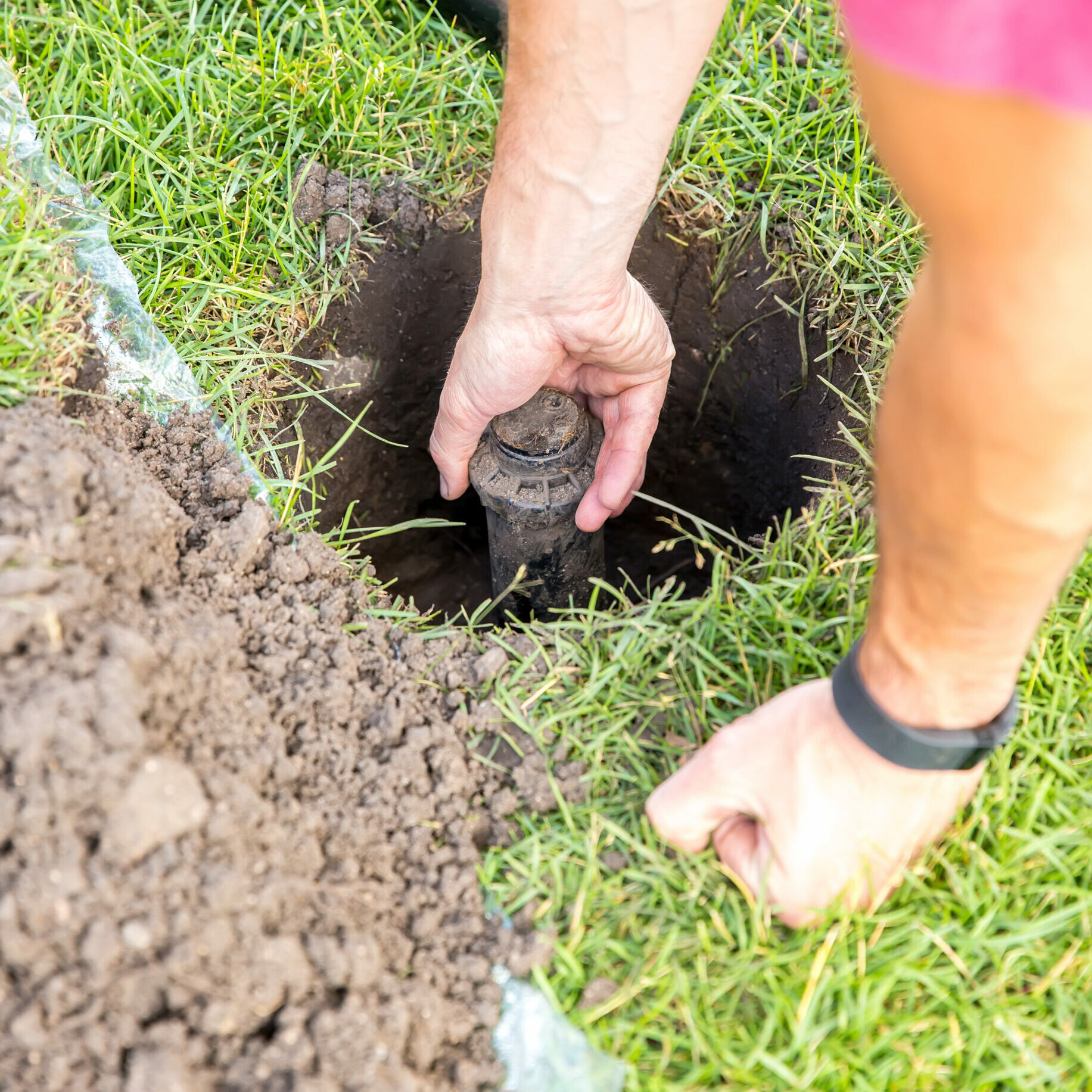 Close-up of a man's hands as he replaces an in-ground sprinkler head. In this image he is taking out the old sprinkler head and has a new one laying on the ground near the top of the image.